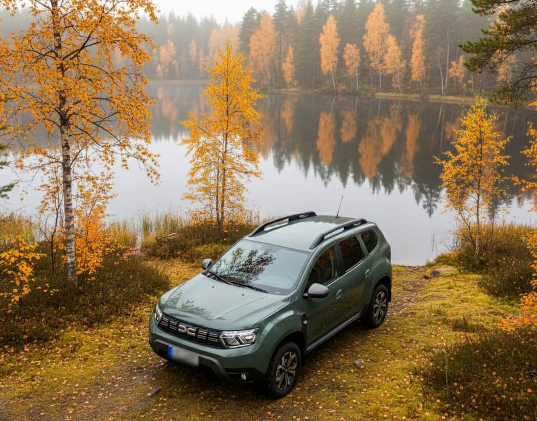 Dacia Duster parked by a lake with autumn trees in the background.
